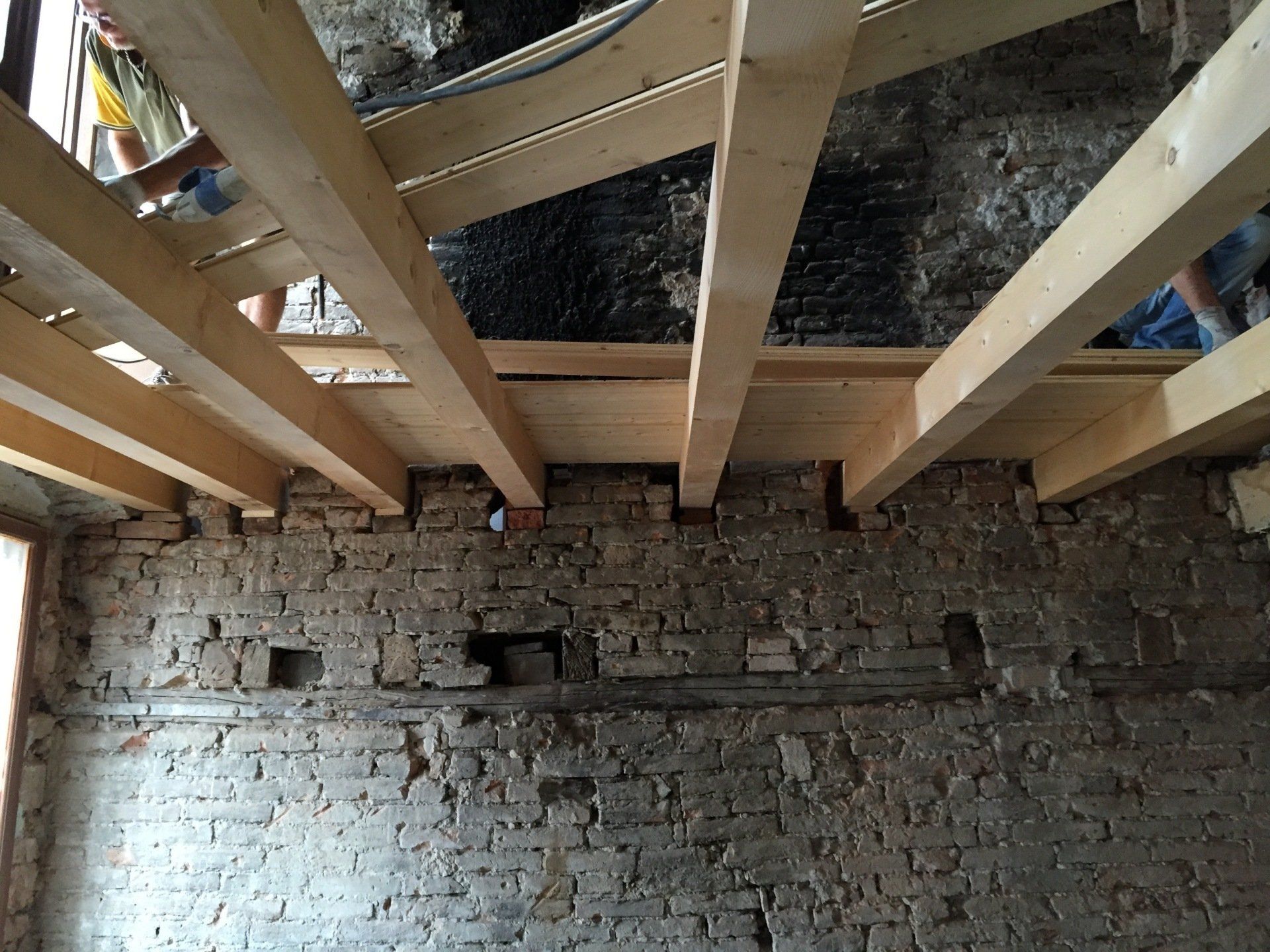 Travi di legno a vista sopra una parete di pietra grezza in un soffitto interno non rifinito.