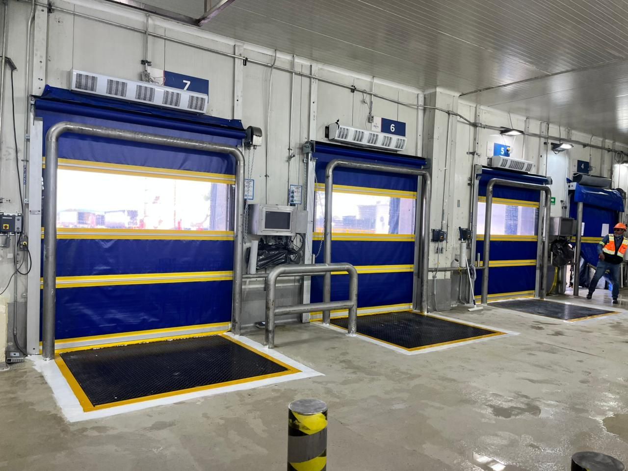 Tres puertas industriales azules y amarillas etiquetadas con los números 5 y 7, cada una con una plataforma, dentro de un edificio blanco.