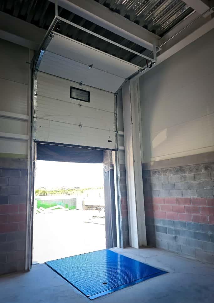 Vista interior de una puerta de muelle de carga abierta con una placa metálica azul sobre el suelo de hormigón.
