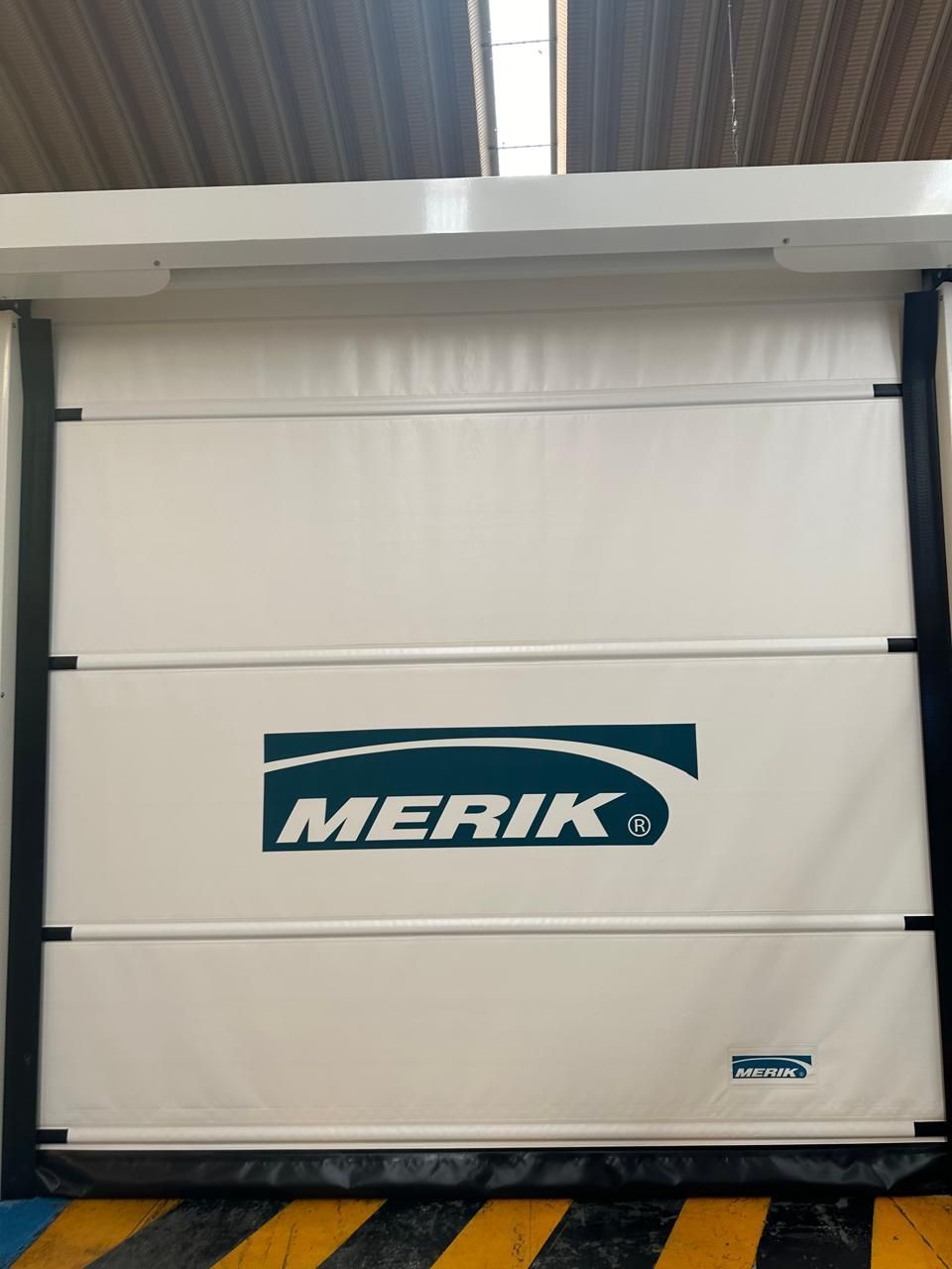 Puerta industrial Merik blanca con detalles y logotipo en negro, en un entorno de fábrica.