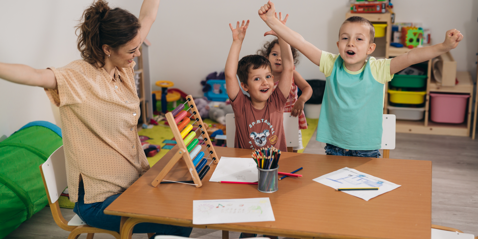 A group of children are sitting at a table with their arms in the air.