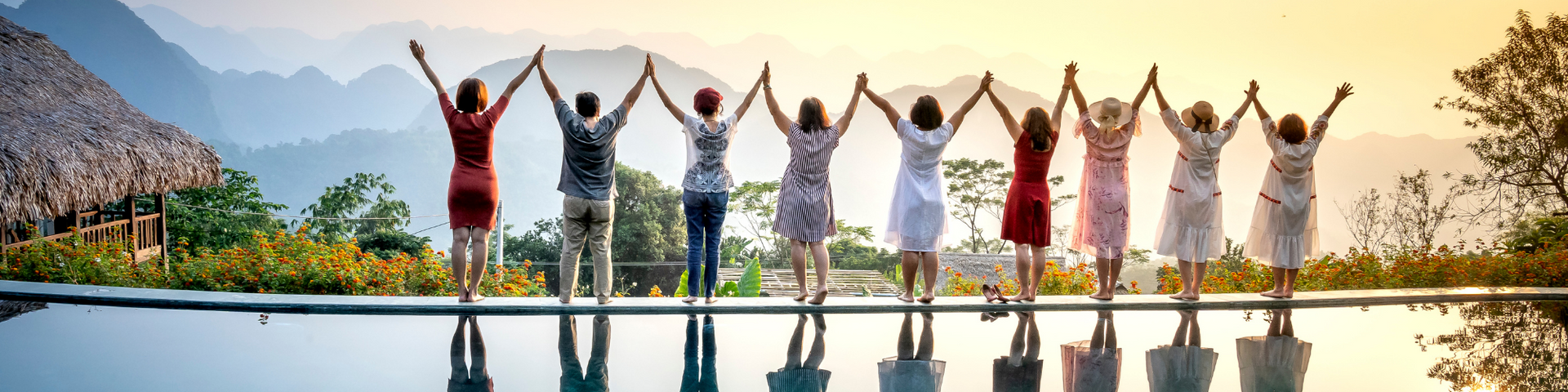 Eight people holding hands and raising them on a pool edge at sunset, with mountains and water behind them