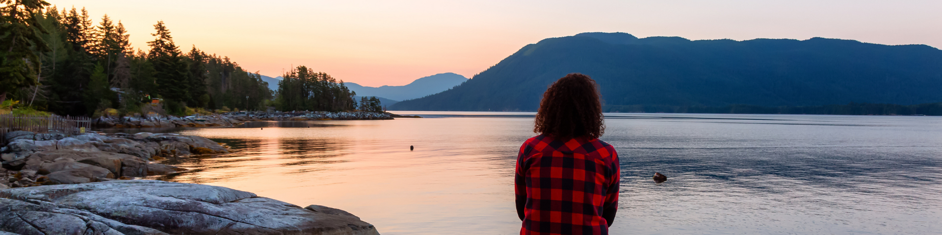 Person in a red plaid shirt sits by a calm lake at sunset with mountains and trees in the distance