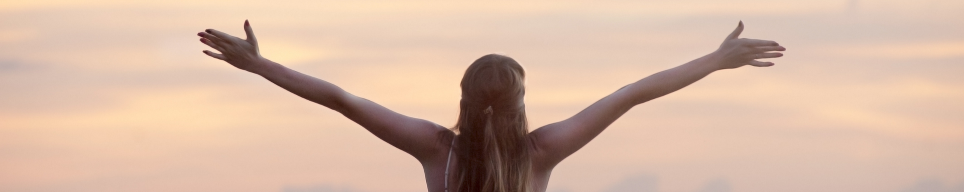 Person standing with arms raised at a beach during sunset, facing the ocean