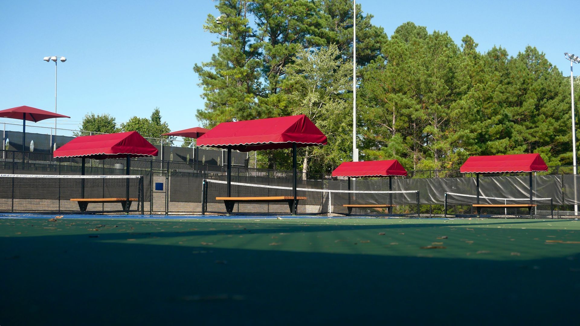 Outdoor tennis courts with red-roofed benches and trees under a clear blue sky