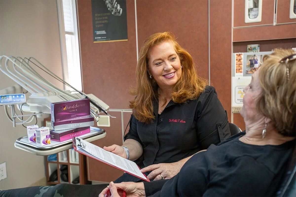 Two women talk at a dental desk; a red-haired staff member holds a clipboard and smiles.