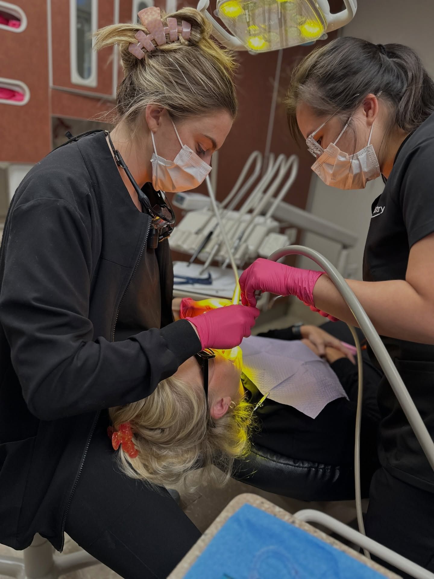 Two dental workers in masks and pink gloves examine a patient in a dental chair with tools nearby