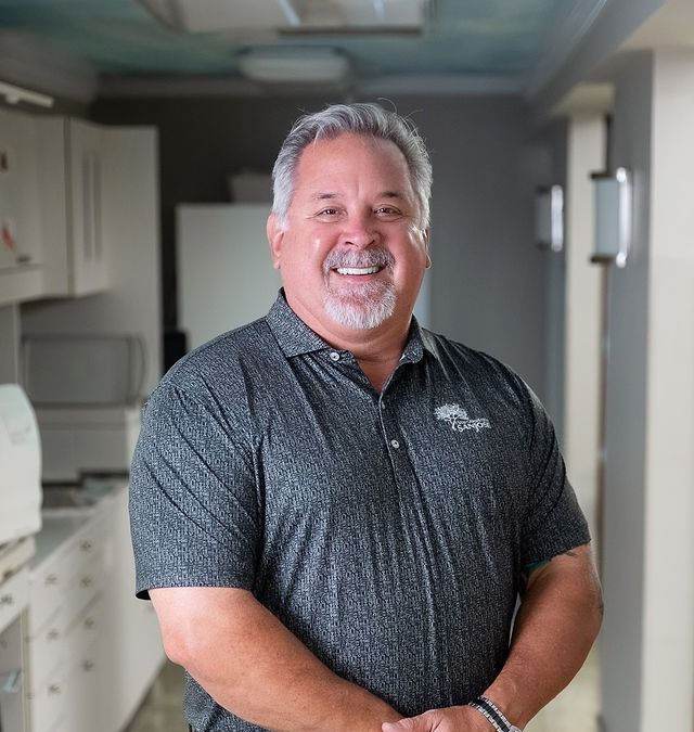 Smiling man in a dark polo shirt standing in a bright kitchen or hallway.