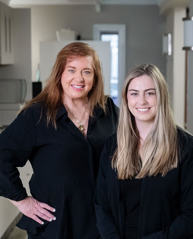 Two women standing indoors, smiling, in black tops near a hallway.