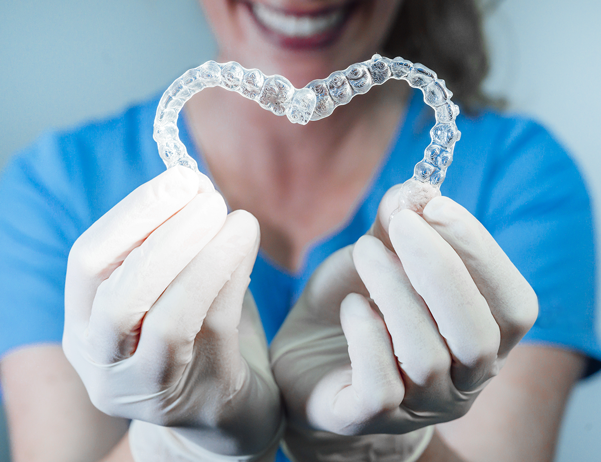 Hands in white gloves holding a clear dental aligner in front of a smiling person