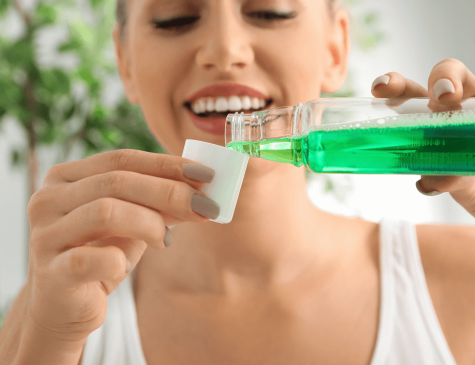 Woman pours green mouthwash into a white cap while smiling in a bright bathroom