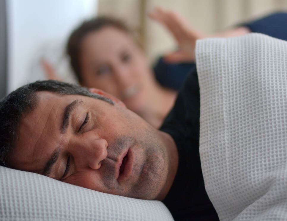 Man sleeping on a pillow under a white blanket, with a blurred person in the background raising a hand