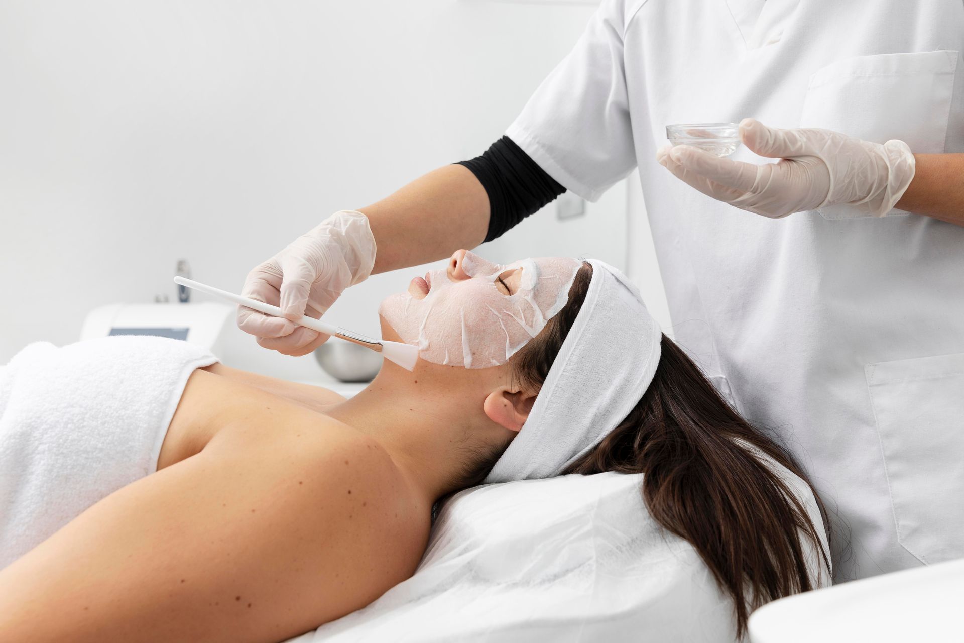 A skincare specialist using a brush to apply a clay mask to a woman’s face in a tranquil spa environment.