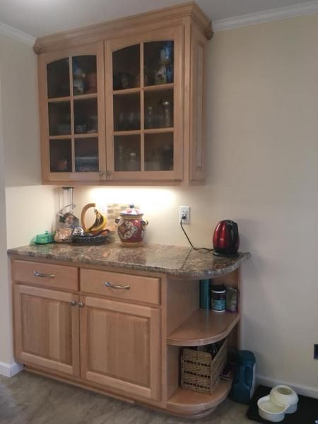 Light-wood kitchen cabinet set against a beige wall, featuring a countertop with items and a glass-front upper cabinet.