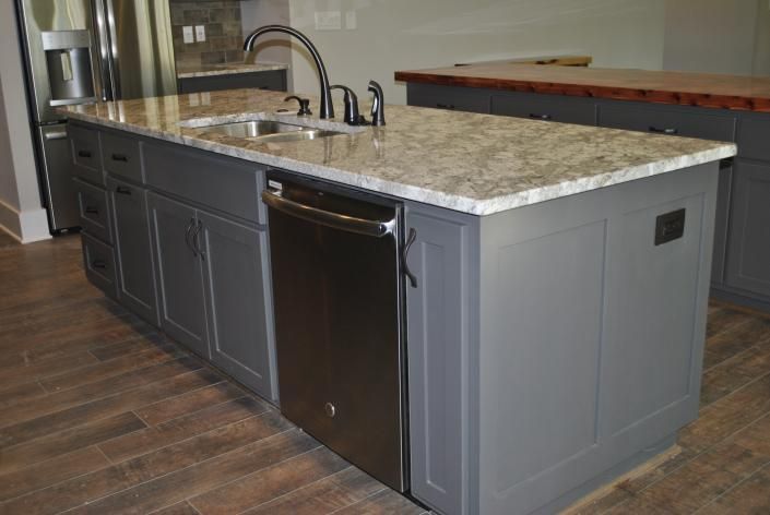 Gray kitchen island with granite countertop, sink, dishwasher, and cabinets on wood-look tile.