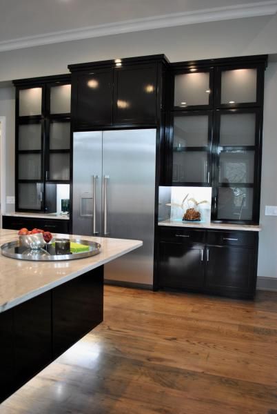 Modern kitchen with black cabinetry, stainless steel refrigerator, and light wood flooring.