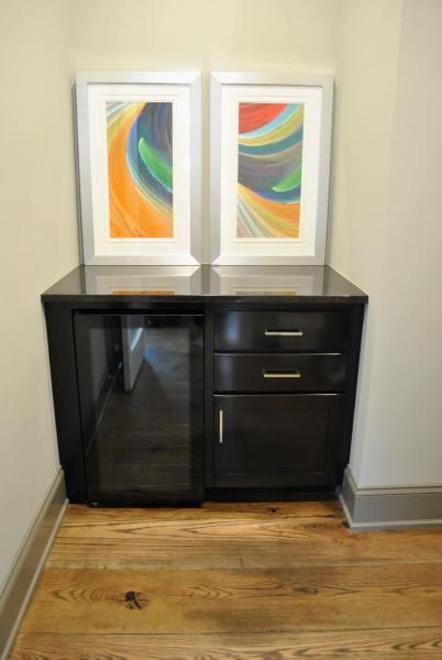 Black cabinet with a mini-fridge and drawers, topped with artwork in a hallway with wood floors.