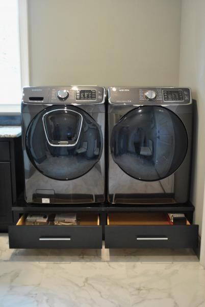 Front-load washer and dryer on black drawers in a laundry room; light marble flooring, gray walls.
