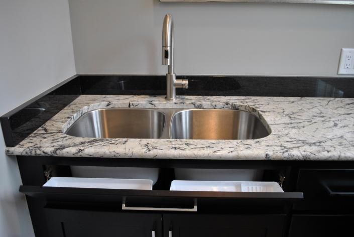 Stainless steel double sink in a kitchen, with dark cabinets and a light-colored countertop. Drawers are open.