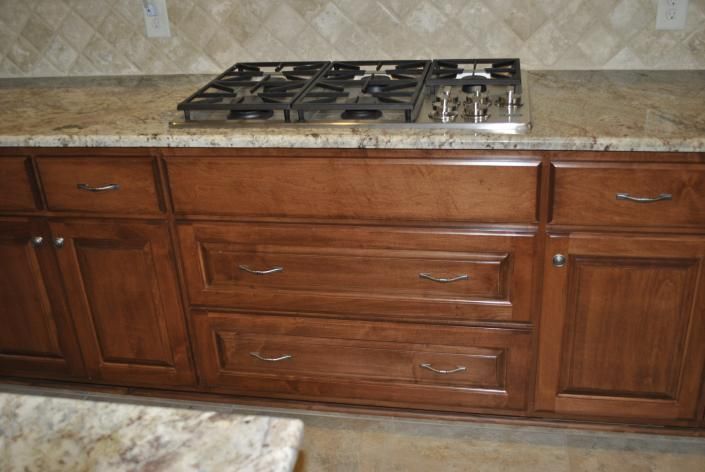 Kitchen cabinets with a gas stovetop on a granite countertop. Brown cabinets, tan backsplash.