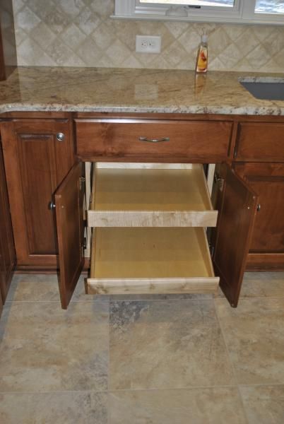 Kitchen cabinet with open doors revealing pull-out shelves. Brown wood, tan countertop, and tile floor.