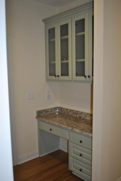 Built-in desk with overhead cabinets in a room. Light green cabinets, granite countertop, drawers, and wooden floor.