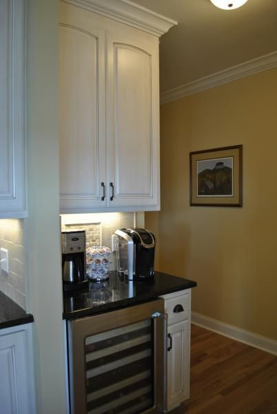 Kitchen coffee station with white cabinets, black countertop, wine fridge, and yellow wall.