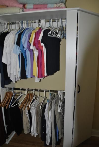 Closet with hanging clothes on rods. White cabinet, neutral wall. Various colored shirts and pants visible.