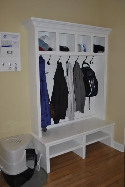 White built-in entryway bench with hooks for coats and upper storage compartments against a light yellow wall.