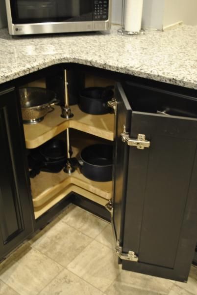 Corner kitchen cabinet with rotating shelves holding cookware. Dark cabinets, granite countertop, light floor.