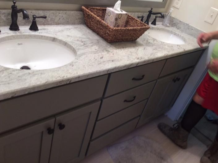 Bathroom vanity with two sinks, gray cabinets, granite countertop, and a person.