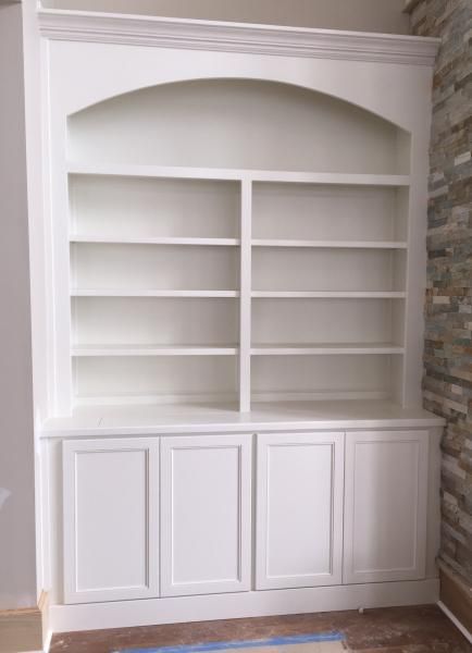White built-in bookshelf with arched top and cabinet doors, against a stone wall.