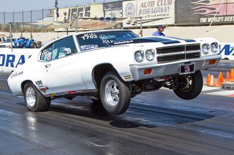 A white classic muscle car performing a wheelie on a drag racing track with a stadium background.