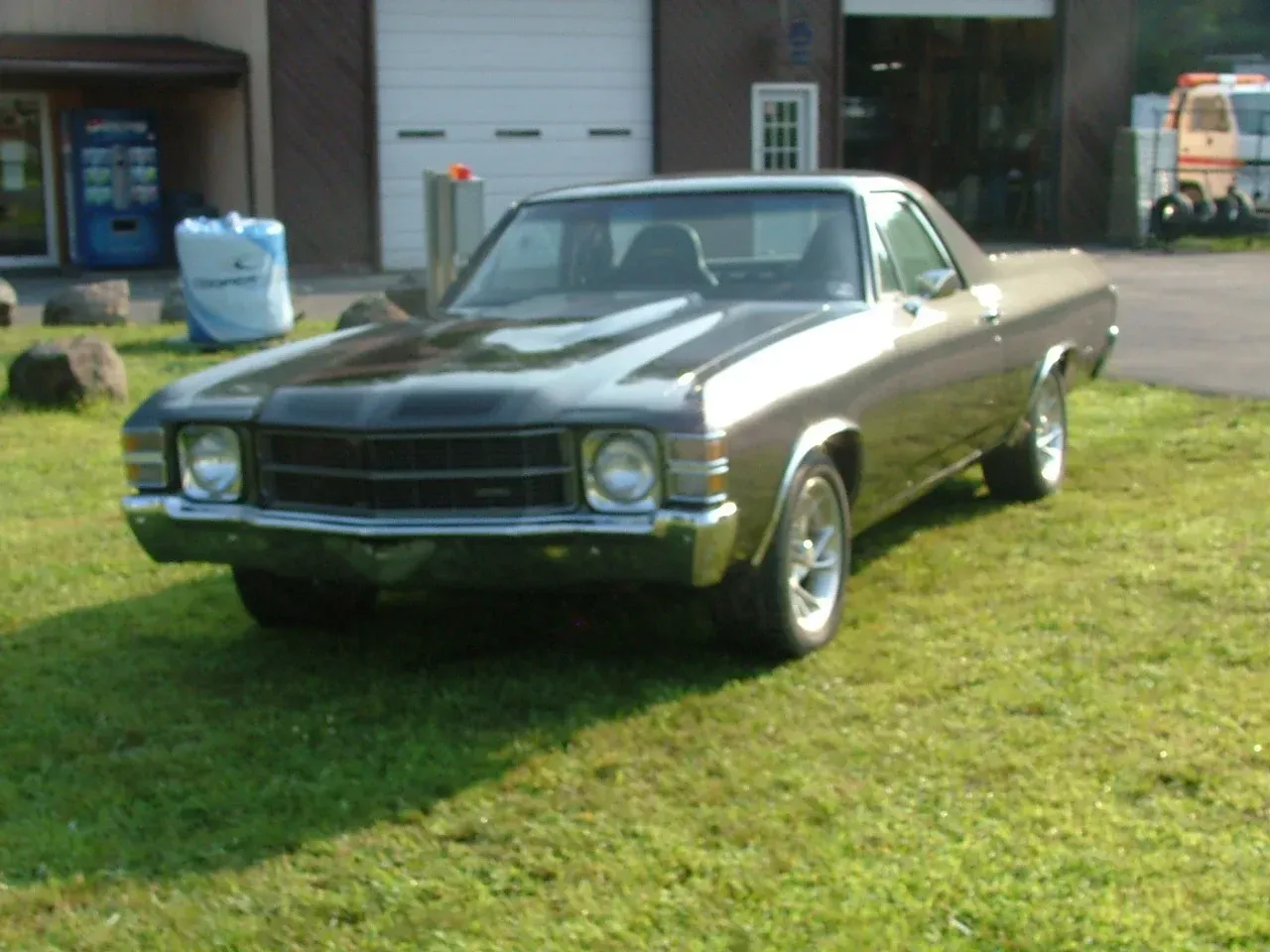 A tan Chevrolet El Camino parked on a grass lawn in front of a building with a large white garage door.