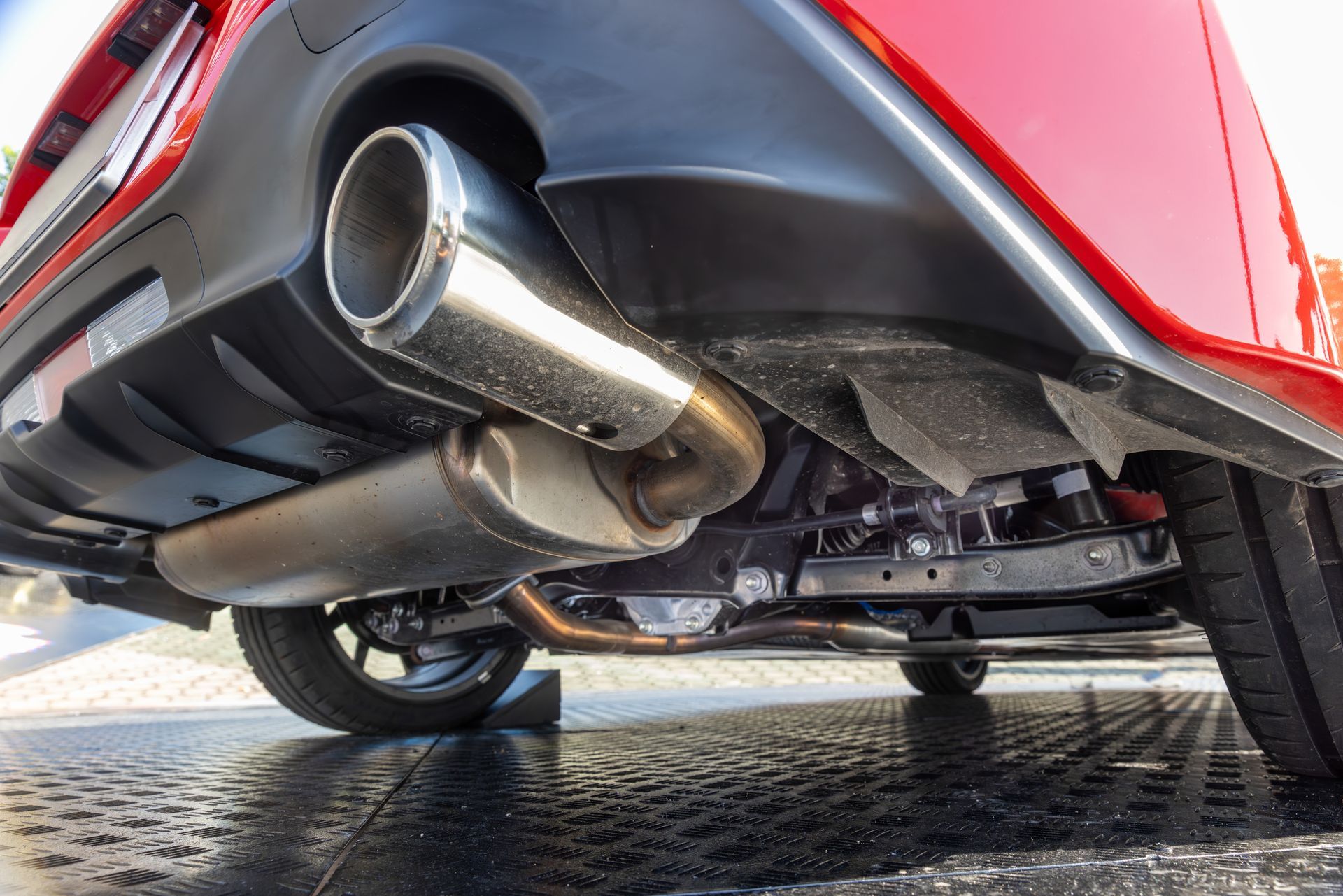 A low-angle view of the undercarriage and shiny exhaust pipe of a red car parked on a textured surface.