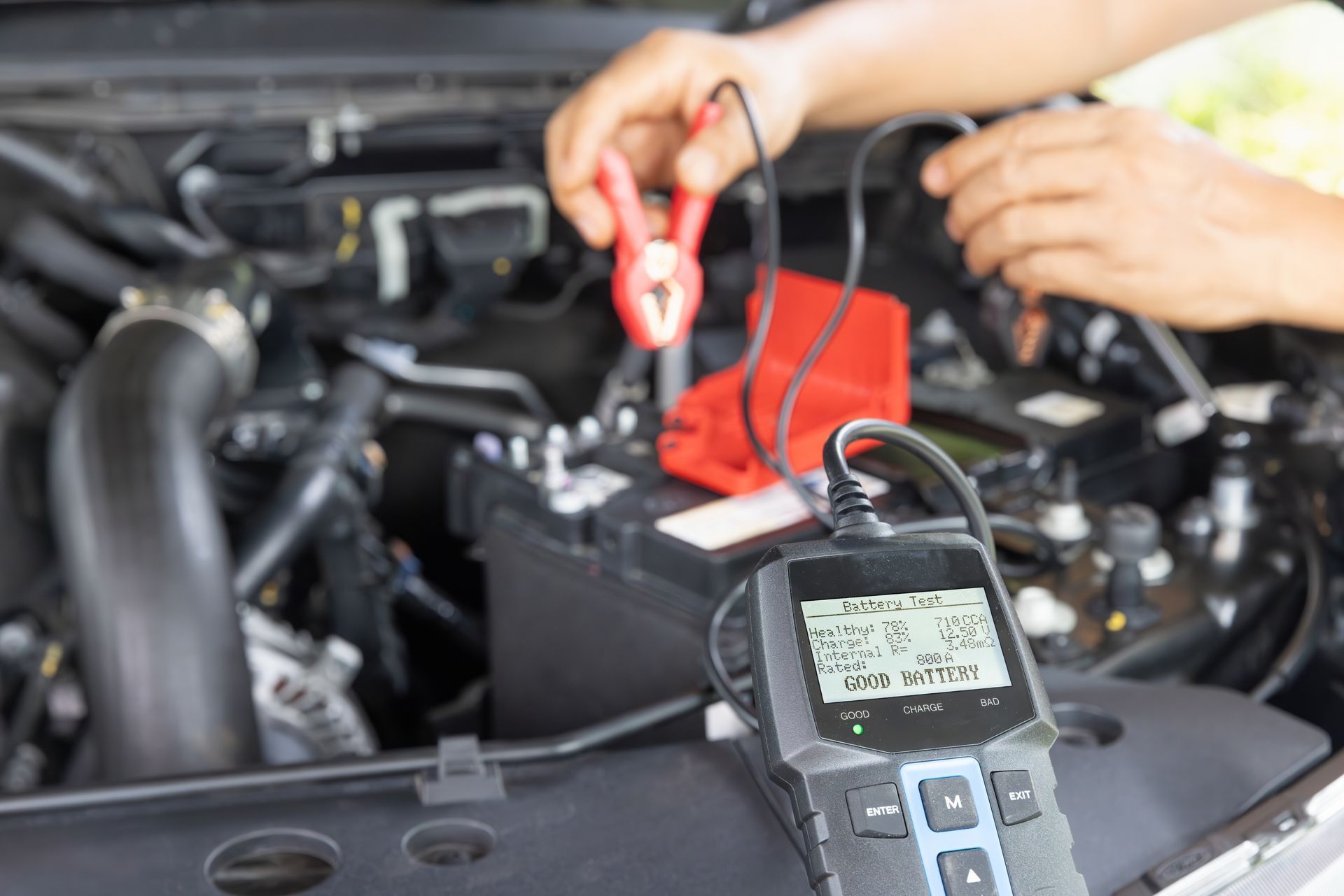 A person uses a digital battery tester to check the charge of a car battery under the hood of a vehicle.