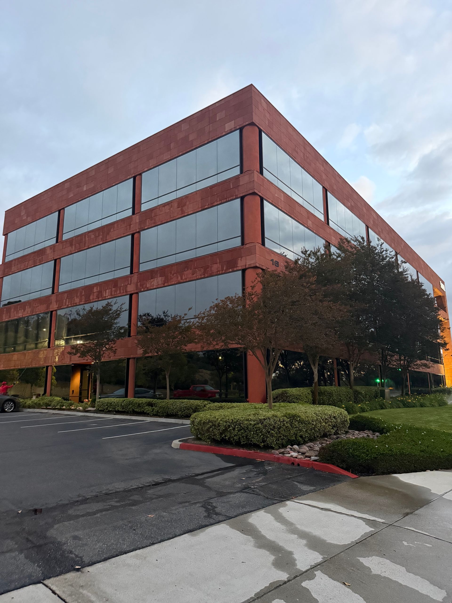 Brick office building with reflective windows beside a parking lot and landscaped trees under a cloudy sky