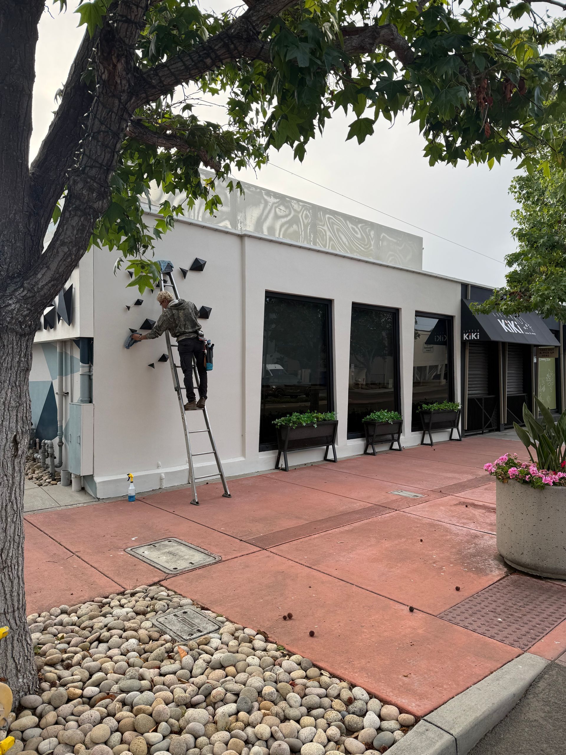 White building with black-framed windows, red patio, potted flowers, and a tree in the foreground.