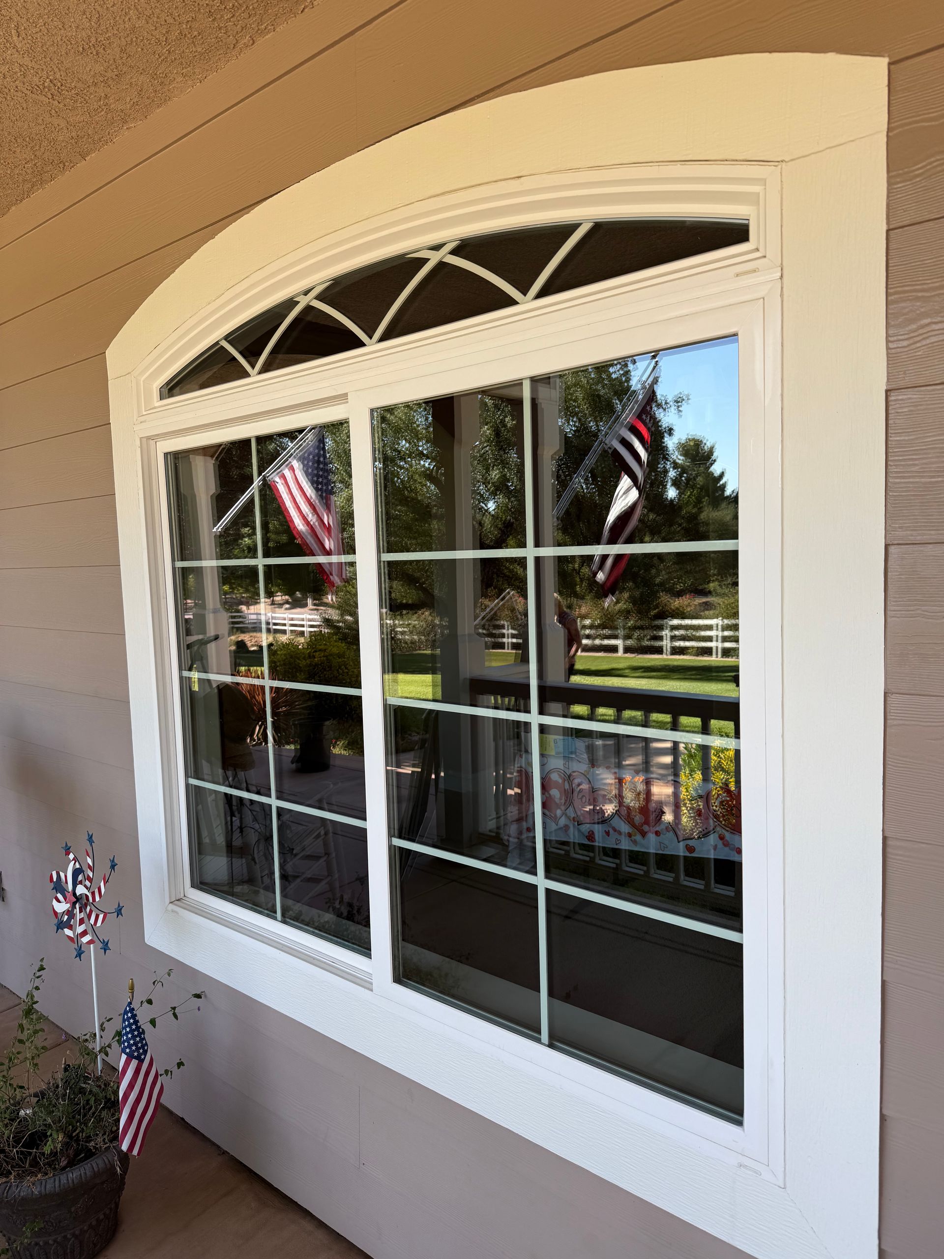 White-trimmed arched window with grid panes reflecting an American flag and porch railing on a beige wall