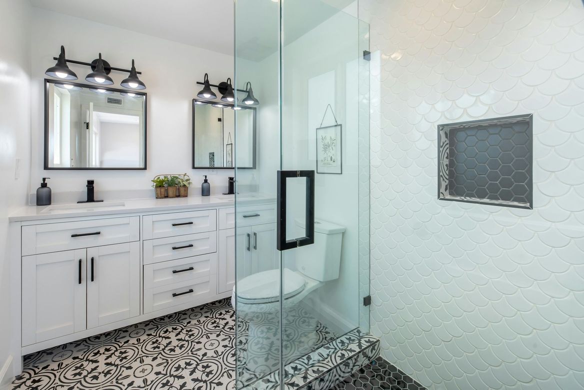 Modern white bathroom with patterned floor, double vanity, glass shower, and black fixtures.