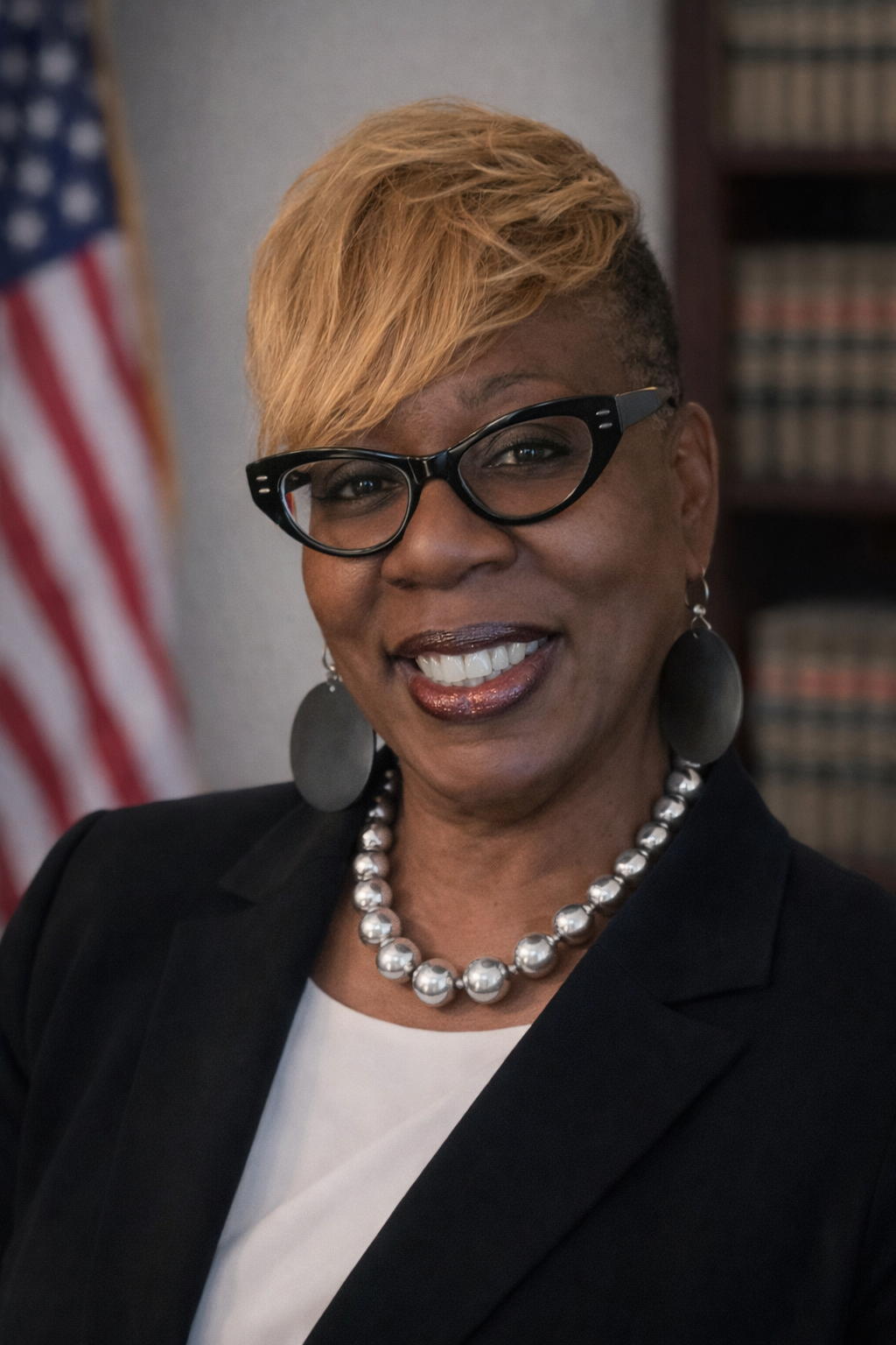 Woman with glasses, blazer, pearl necklace, smiling, in front of an American flag and bookshelves.