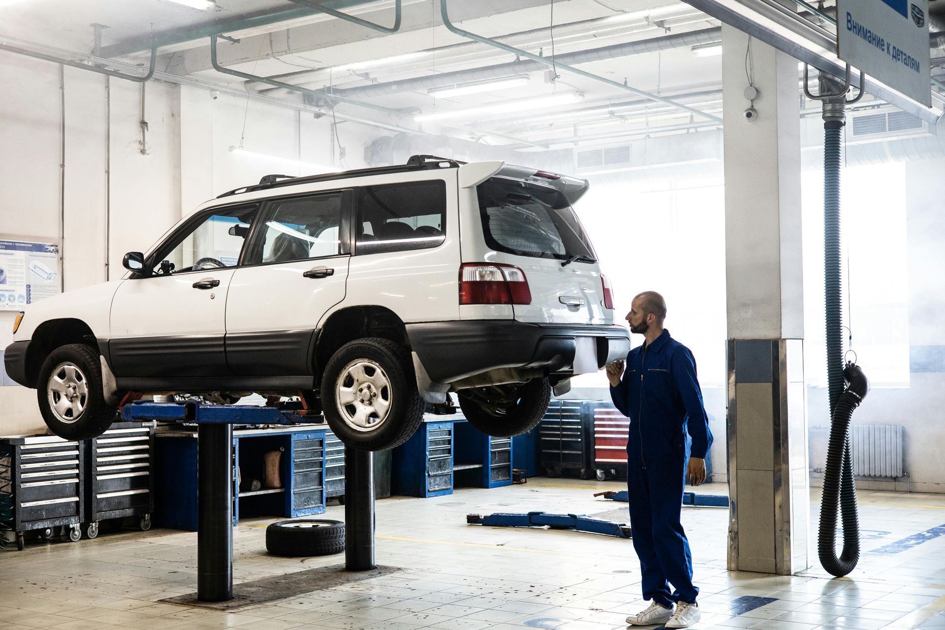 A mechanic in blue coveralls examines a white SUV raised on a lift inside an auto repair shop.