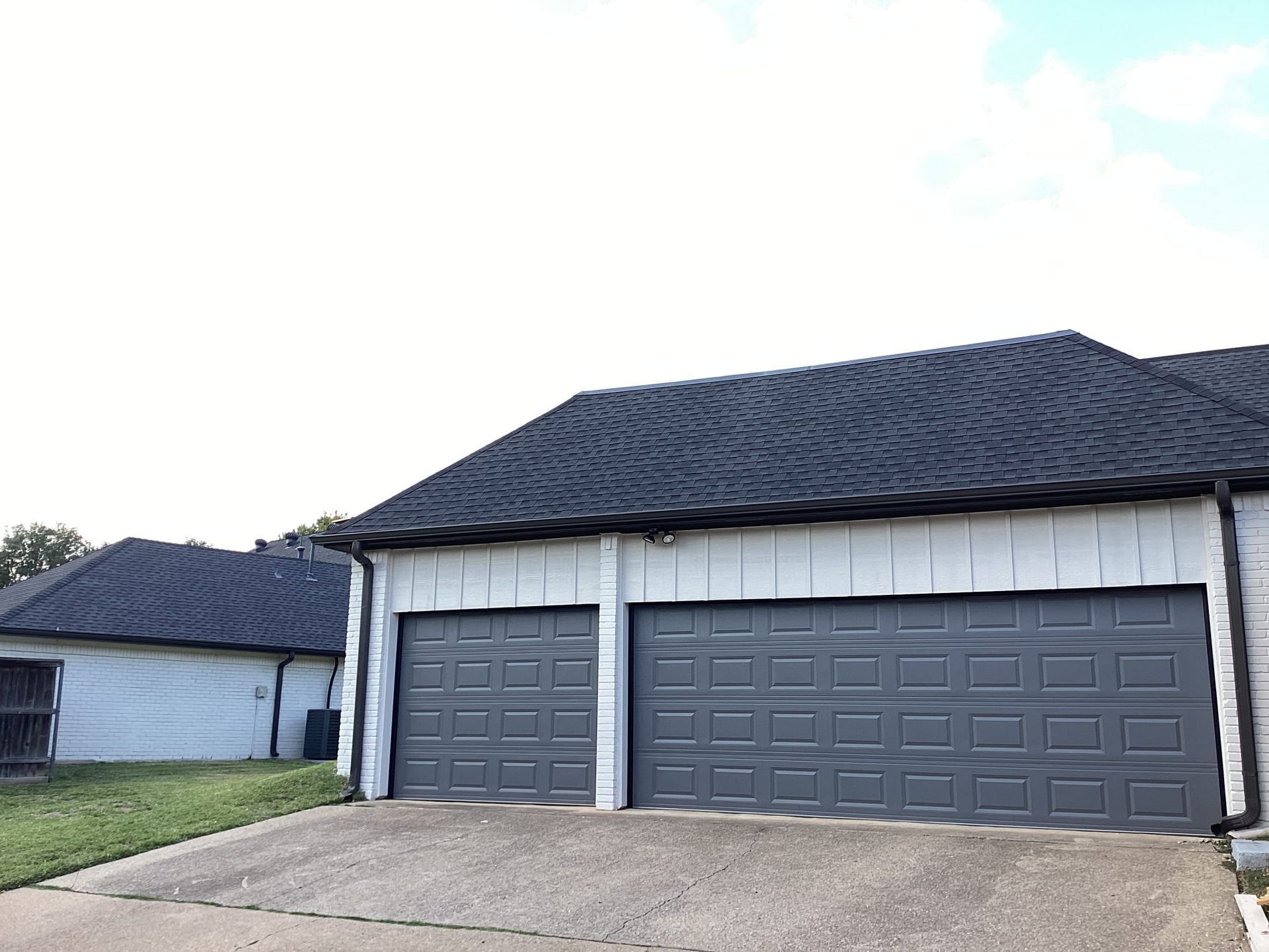 Two gray garage doors on a house with a dark shingled roof and white vertical siding, viewed from a paved driveway.