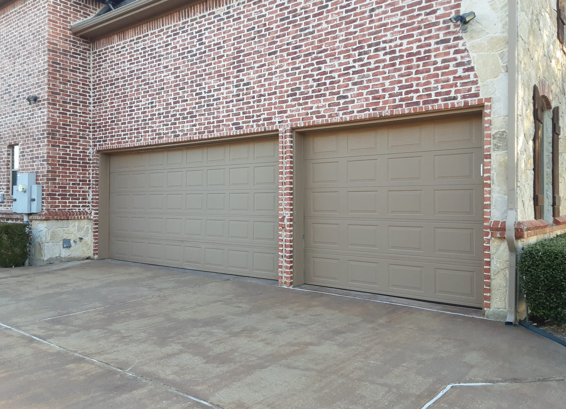 Two beige garage doors on a brick building with a concrete driveway.