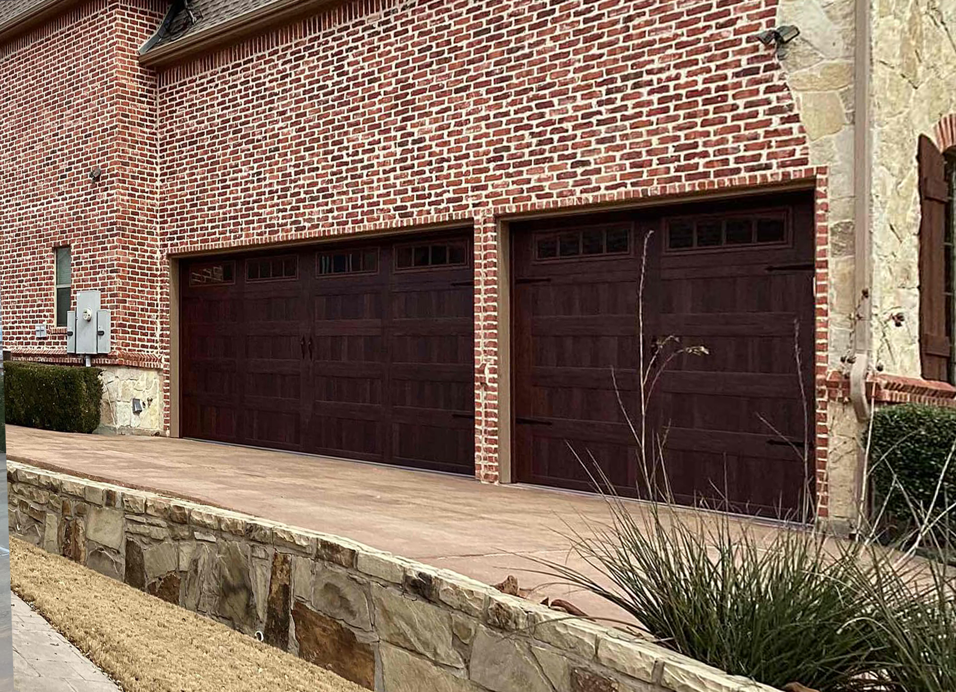 Two dark brown garage doors on a brick building with a concrete driveway and landscaping.