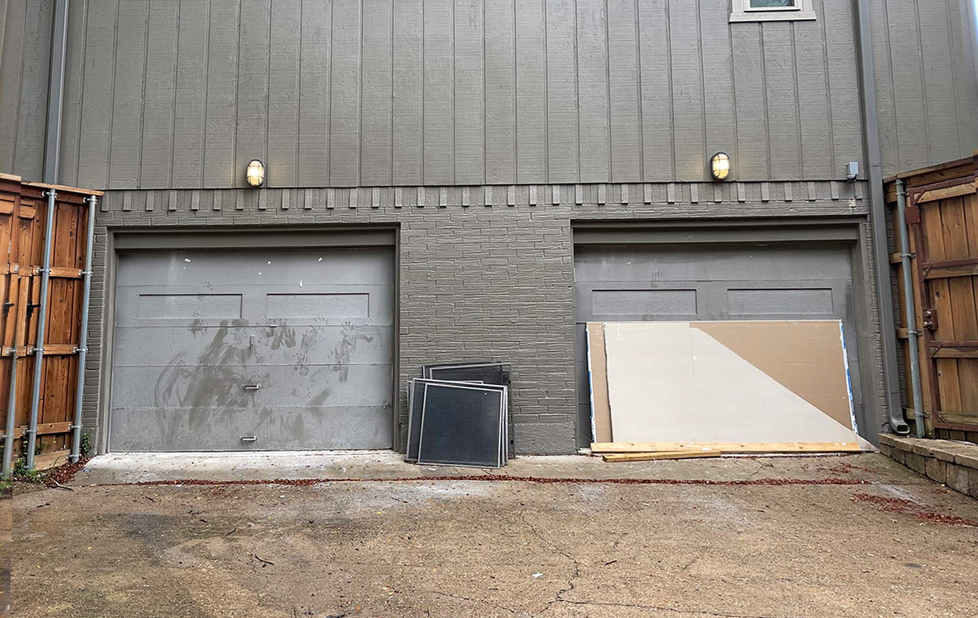 Two gray garage doors under a gray building, flanked by wooden fences. Gravel driveway.