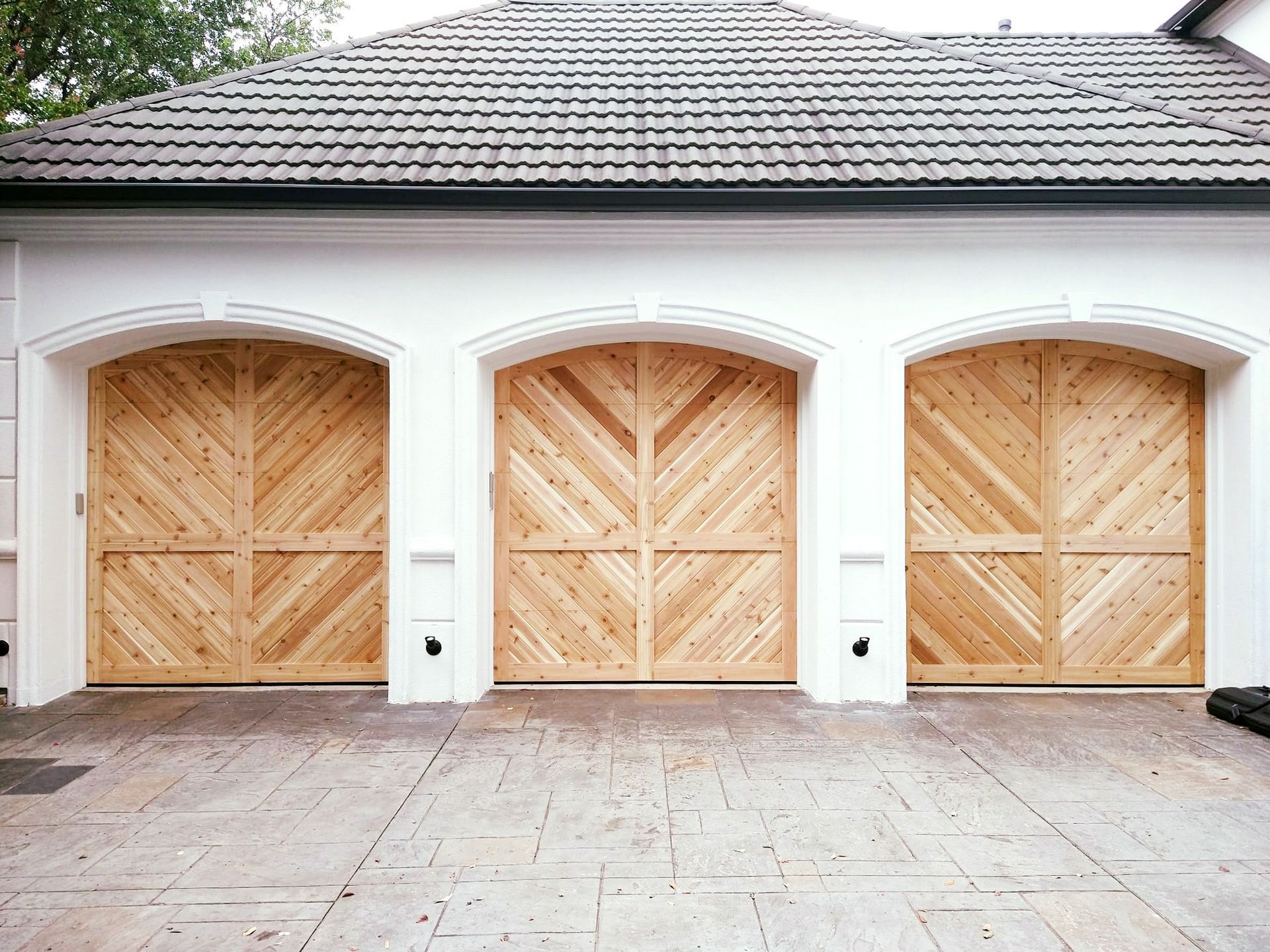 Three wooden arched garage doors with a herringbone pattern set in a white wall with a shingled roof above.