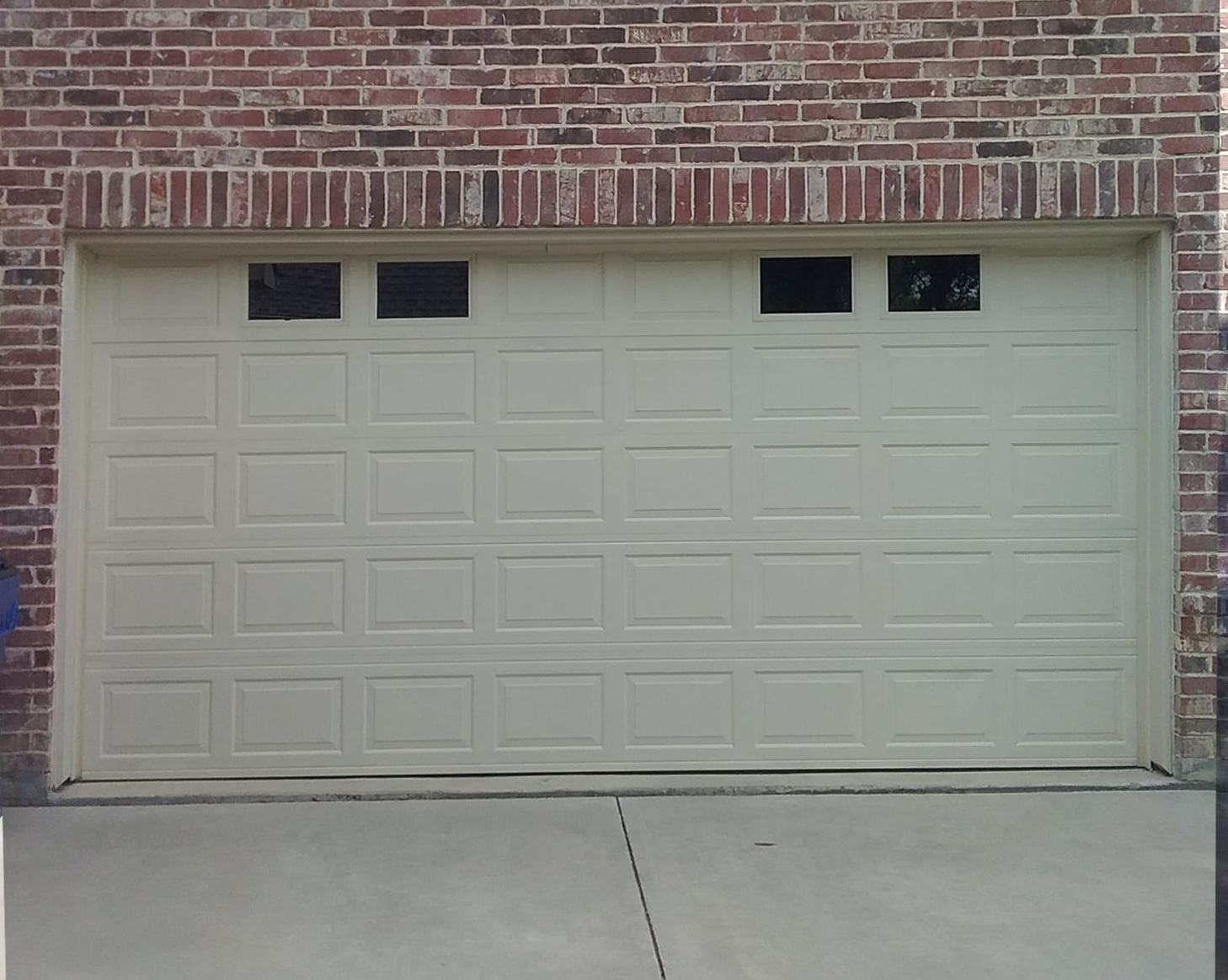 Beige garage door with square windows, set against a red brick wall and concrete driveway.