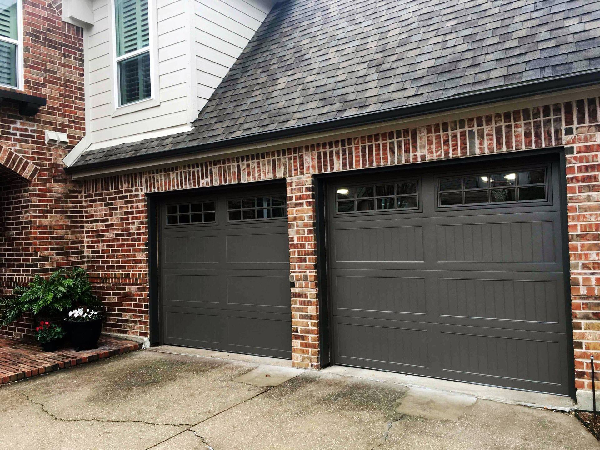 A residential house exterior featuring a two-car garage with dark grey doors and brick siding under a shingled roof.
