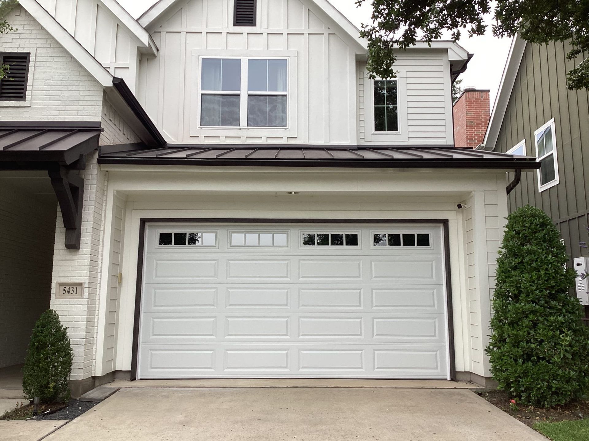 A two-story white suburban house featuring a metal roof, a prominent garage door with windows, and green landscaping.